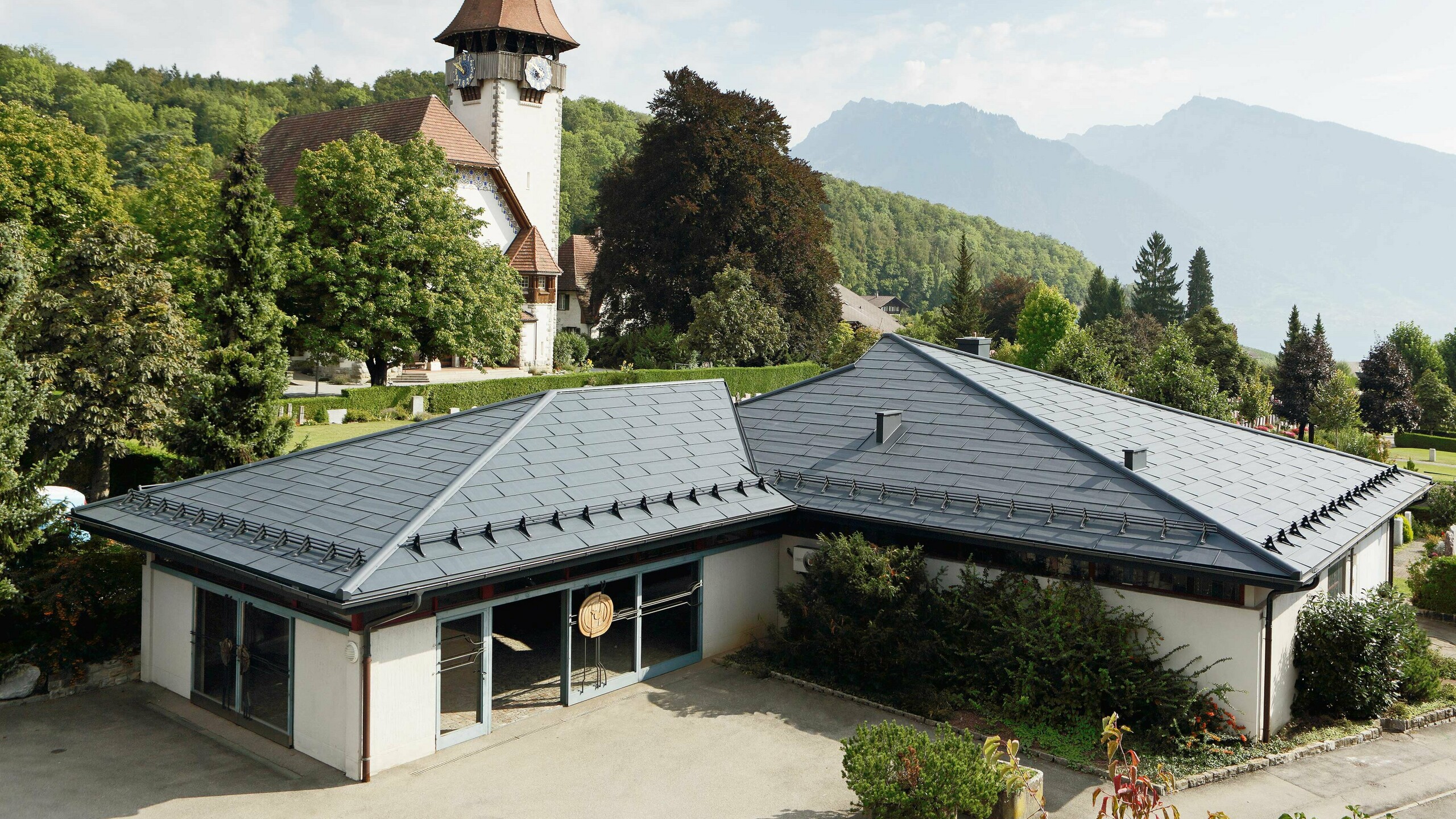 Casa funeraria con pannelli FX.12 PREFA in colore antracite, con una chiesa sullo sfondo e il cimitero accanto.