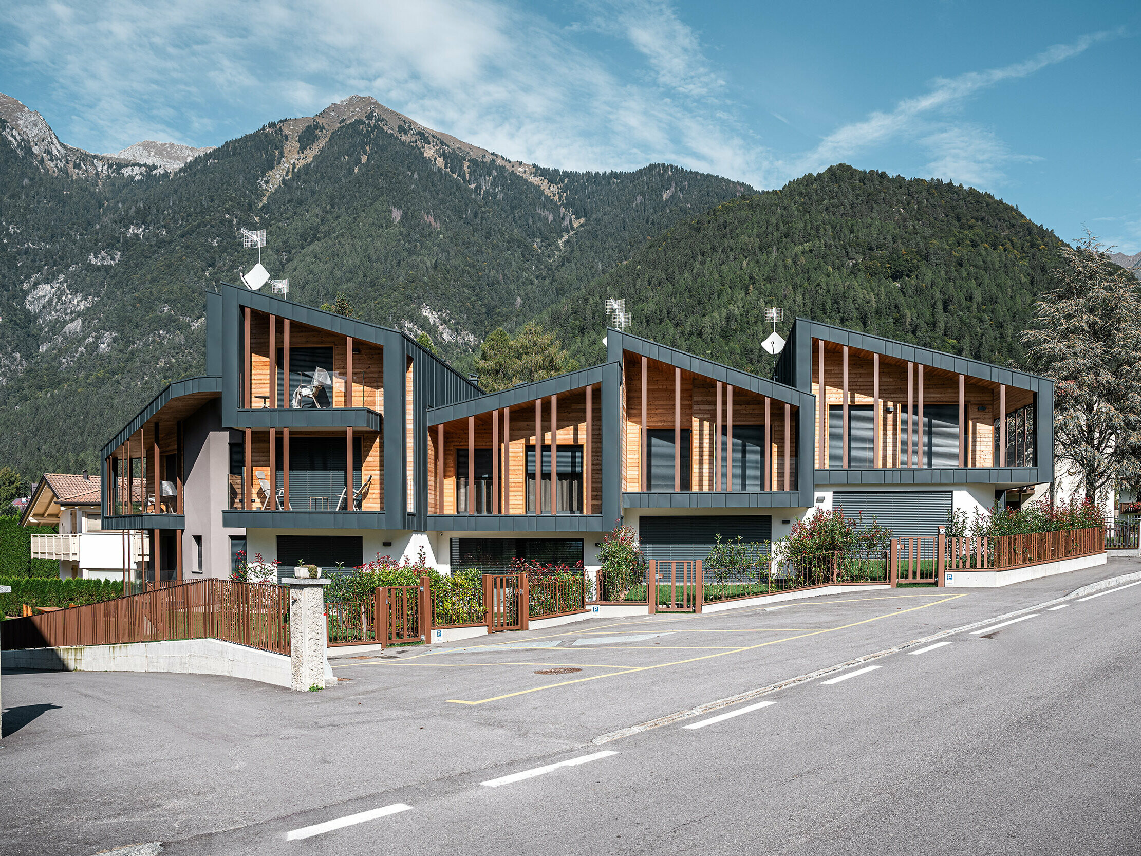 Moderni chalet per vacanze con tetti in metallo Prefalz di color antracite ed elementi in legno in una valle alpina, accanto ad una strada con giardini antistanti recintati e un impressionante panorama montuoso sullo sfondo.