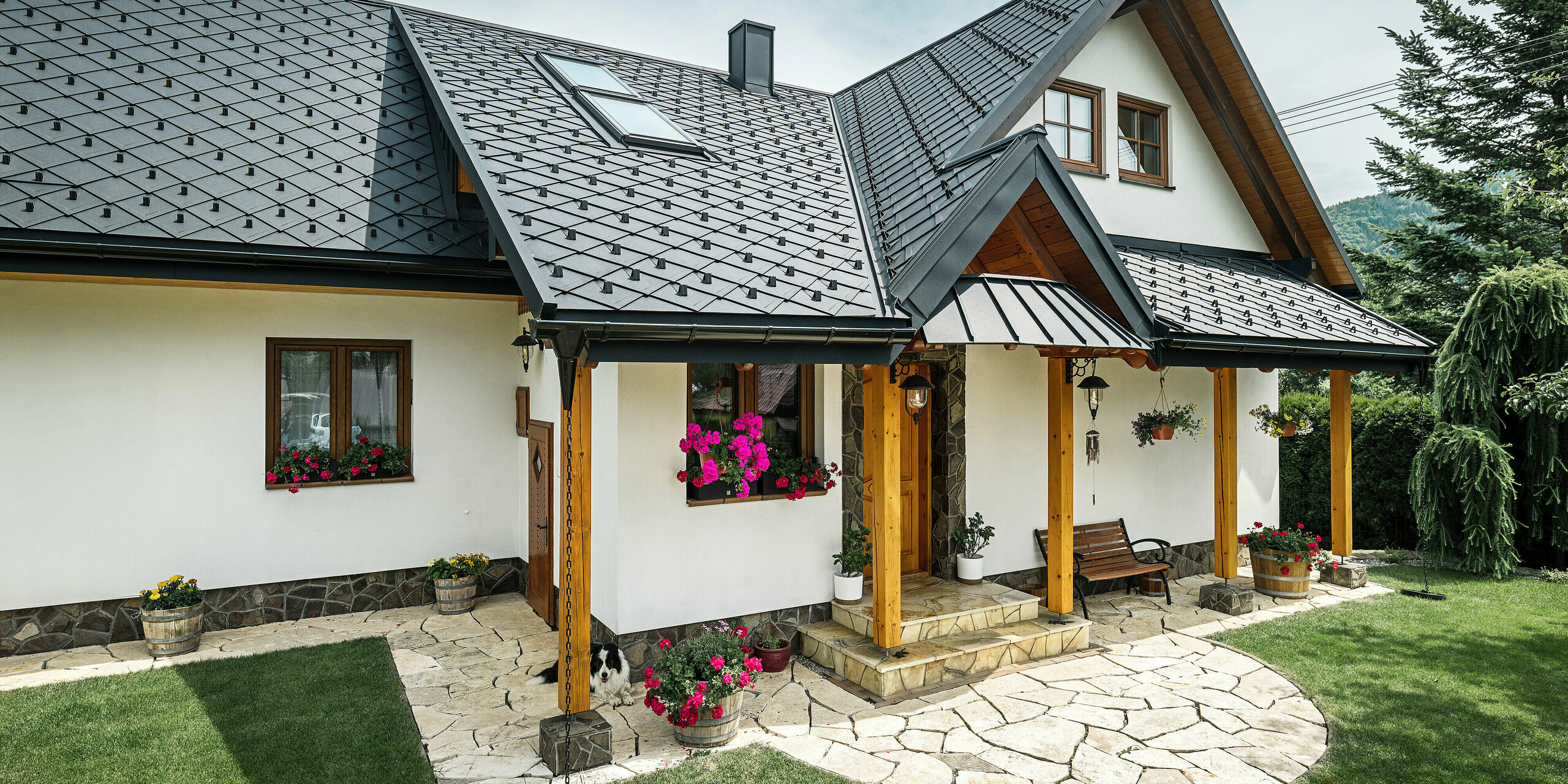 Eingangsbereich eines traditionellen Einfamilienhauses in Stara Bystrica, Slowakei, mit einem Dach aus PREFA Dachrauten 29 × 29 in P.10 Schwarz. Das Aluminiumdach in Schuppenoptik umfasst Dachschrägenfenster und ein Vordach, das mit PREFALZ verkleidet ist. Der Eingangsbereich wird durch hölzerne Stützen und eine Naturstein-Terrasse akzentuiert. Farbige Blumenarrangements und eine gepflegte Grünfläche verleihen dem Haus ein einladendes und charmantes Erscheinungsbild. Der liegende Border Collie vor dem Haus bewacht die Eingangstüre. Der ruhende Hund macht das Bild vom konventionellen Familienleben in ländlicher Umgebung perfekt.