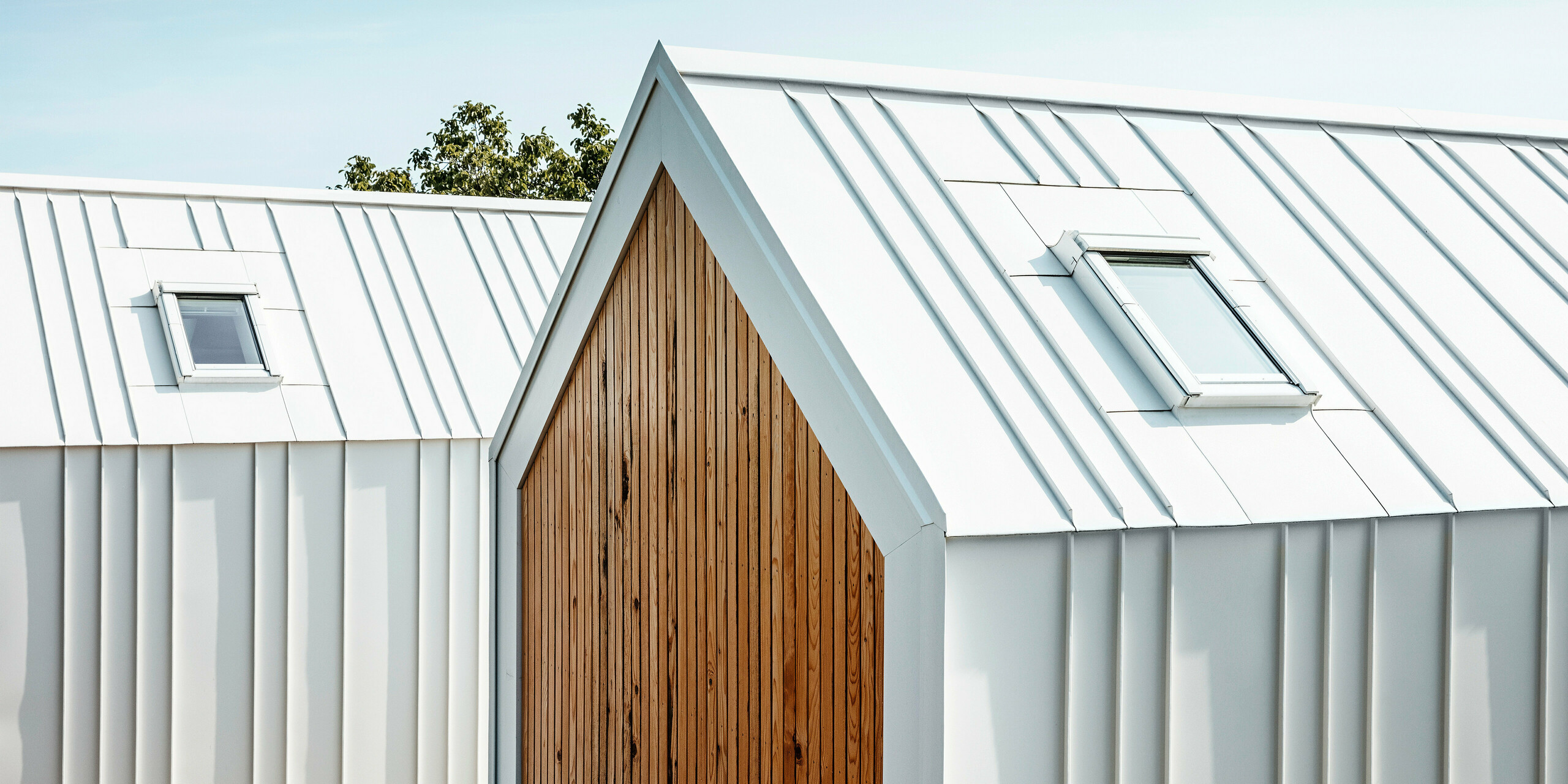 Detailansicht der weißen PREFALZ Dach- und Fassadenverkleidung der Tiny Häuser 'Pri Momi' in Kančevci, Slowenien. Die Gebäude sind in einer modernen Kombination aus hochwertigem Aluminium in P.10 Reinweiß und naturbelassenem Holz gestaltet. Das Bild zeigt die präzise ausgeführten Falze und den eleganten Übergang vom Dach zur Fassade, der ohne sichtbare Unterbrechung der vertikalen Stehfalze realisiert wurde. Die harmonische Materialwahl unterstreicht das minimalistische Design der exklusiven Gästeunterkunft.