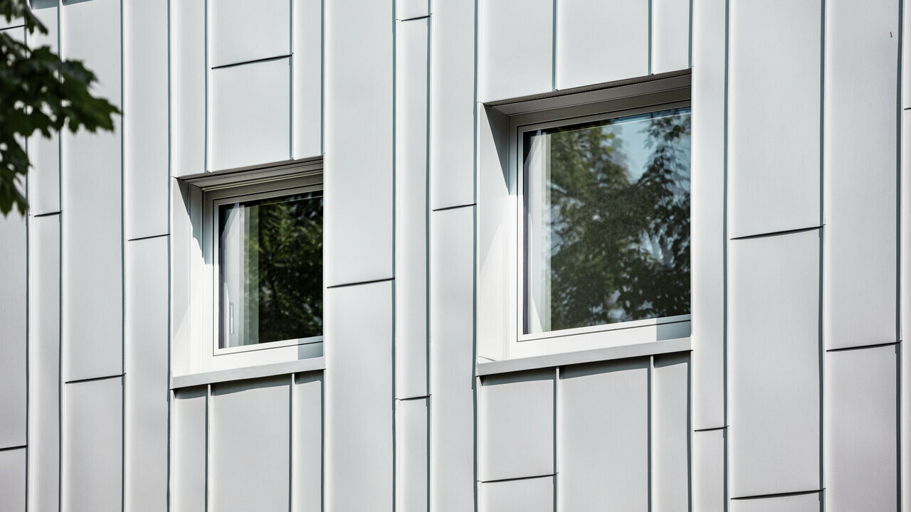 Primo piano della facciata di un edificio moderno con lastre verticali in alluminio Prefalz color silver metallizzato. Due finestre rettangolari con cornici scure riflettono il verde degli alberi e spezzano il disegno strutturato della facciata.
