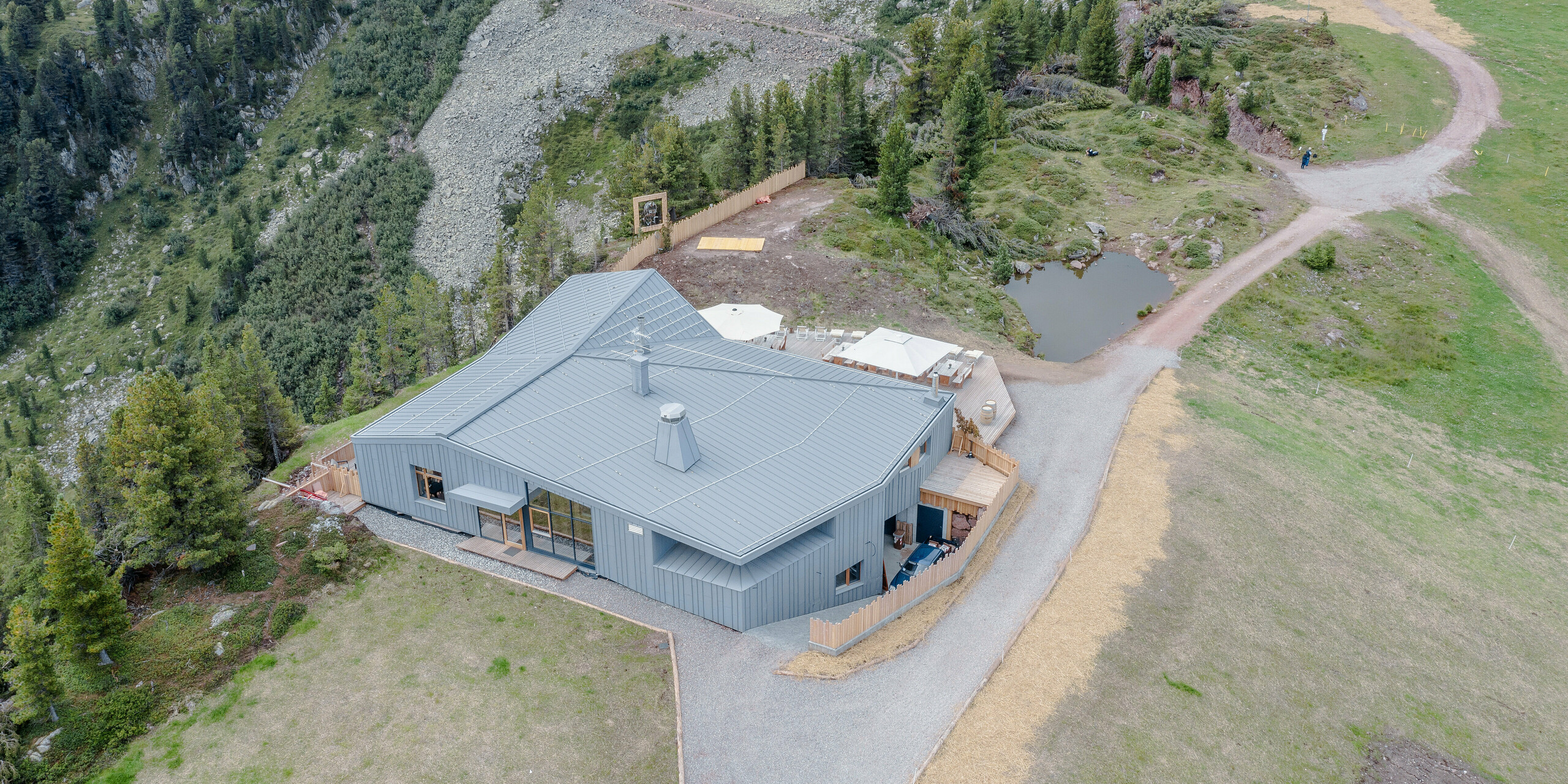 Drohnenaufnahme des Ristorante Busabelle, umgeben von einer alpinen Landschaft mit Wanderwegen, die sich durch die grünen Hänge schlängeln. Das Restaurant, mit seiner hellgrauen PREFALZ Dach- und Fassadenverkleidung, steht nahe einem kleinen natürlichen Teich, was einen friedvollen Rückzugsort in der Bergwelt darstellt. Die gestaltete Außenterrasse bietet einen einladenden Speisebereich im Freien, ideal für Gäste, die die reine Bergluft genießen möchten. Die roubste Gebäudehülle aus Blech bietet Schutz vor alpinen Wetterbedingungen.