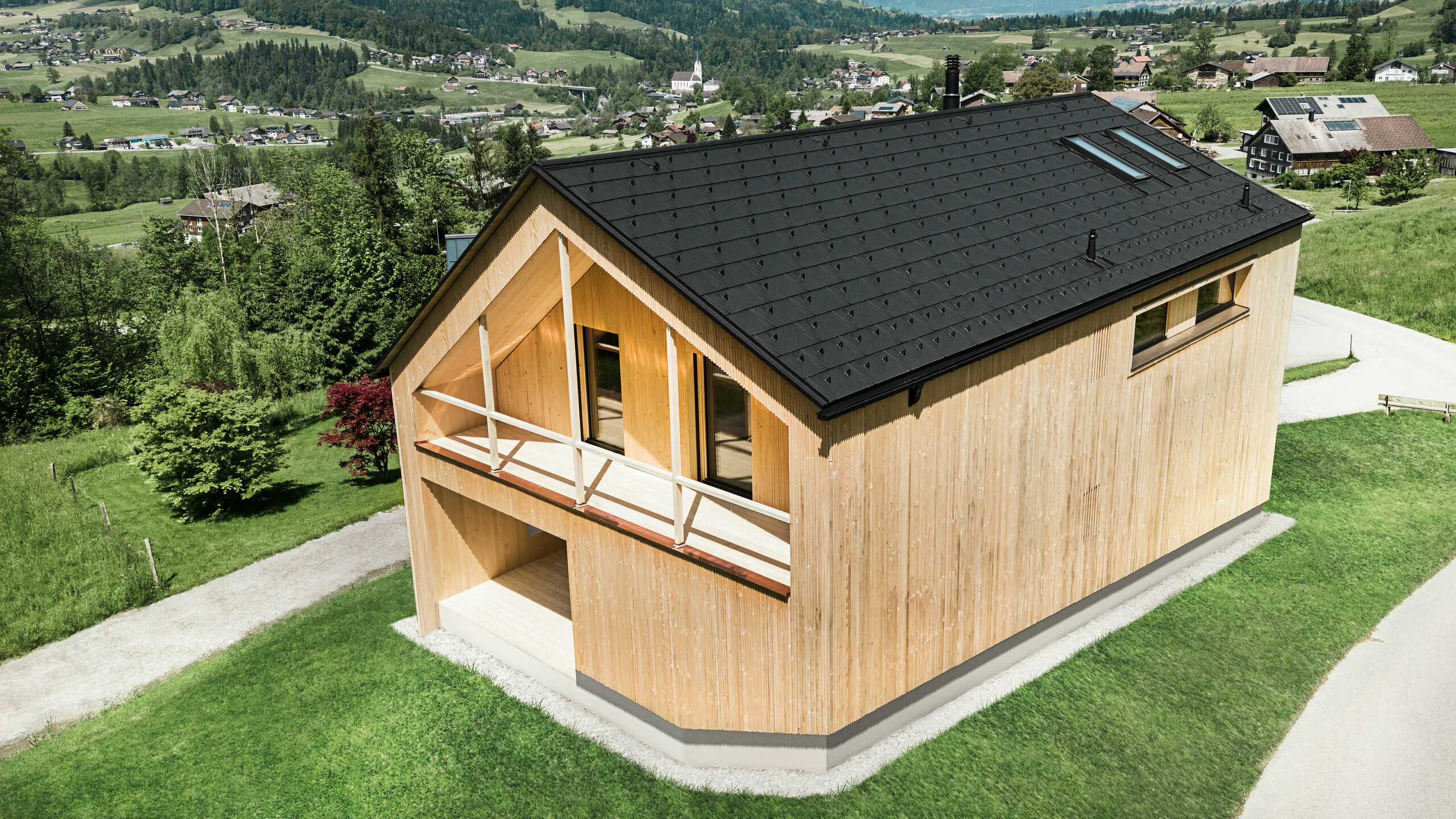 Questa foto mostra una casa vetrina ecologica a Egg, che si staglia sullo sfondo pittoresco di un villaggio austriaco con le sue tegole fotovoltaiche PREFA e le tegole R.16  nere sul tetto. Gli elementi solari sono perfettamente integrati nella struttura del tetto e forniscono energia da una fonte rinnovabile, mentre il legno naturale della facciata e il verde dell'ambiente circostante rappresentano una combinazione di sostenibilità e località. La vista della struttura del villaggio circostante con gli edifici tradizionali sullo sfondo aggiunge un'ulteriore dimensione all'immagine e sottolinea il successo dell'integrazione di metodi di costruzione moderni ed ecologici in paesaggi consolidati.