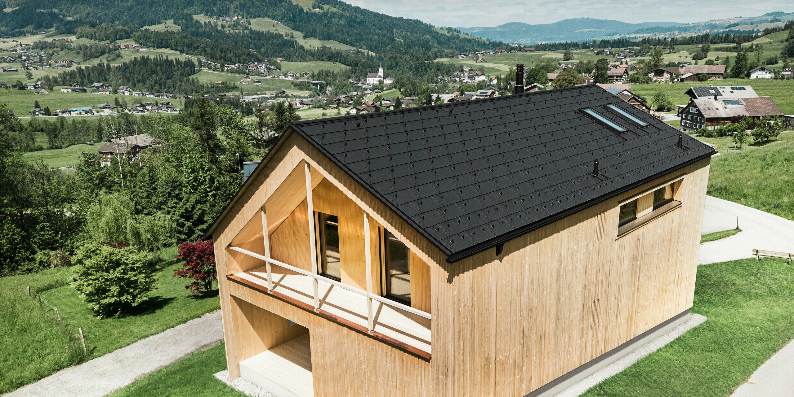 Questa foto mostra una casa vetrina ecologica a Egg, che si staglia sullo sfondo pittoresco di un villaggio austriaco con le sue tegole fotovoltaiche PREFA e le tegole R.16  nere sul tetto. Gli elementi solari sono perfettamente integrati nella struttura del tetto e forniscono energia da una fonte rinnovabile, mentre il legno naturale della facciata e il verde dell'ambiente circostante rappresentano una combinazione di sostenibilità e località. La vista della struttura del villaggio circostante con gli edifici tradizionali sullo sfondo aggiunge un'ulteriore dimensione all'immagine e sottolinea il successo dell'integrazione di metodi di costruzione moderni ed ecologici in paesaggi consolidati.