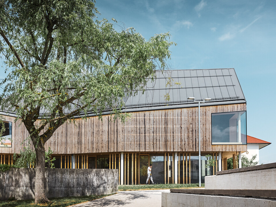 Il nuovo museo palafitticolo con la sua facciata in legno lamellare e la complessa copertura PREFA in grigio scuro vista di lato; un albero incornicia l'edificio nell'angolo in alto a sinistra.