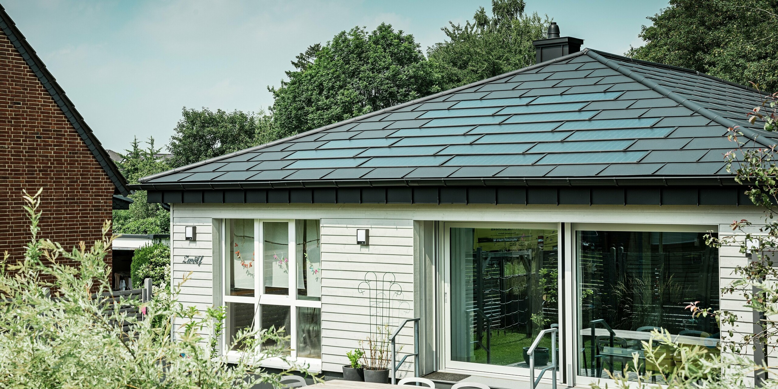 Sanierter Bungalow in Rommerskirchen mit elegantem PREFA Aluminiumdach. PREFA Solardachplatten und Dachplatten R.16 in P.10 Schwarz schützen dauerhaft und erzeugen nachhaltig Strom aus Sonnenenergie. Die großzügige Fensterfront sorgt für viel Licht, die Dachentwässerung mit Saumrinne und Ablaufrohr verbindet Funktionalität und Design perfekt.