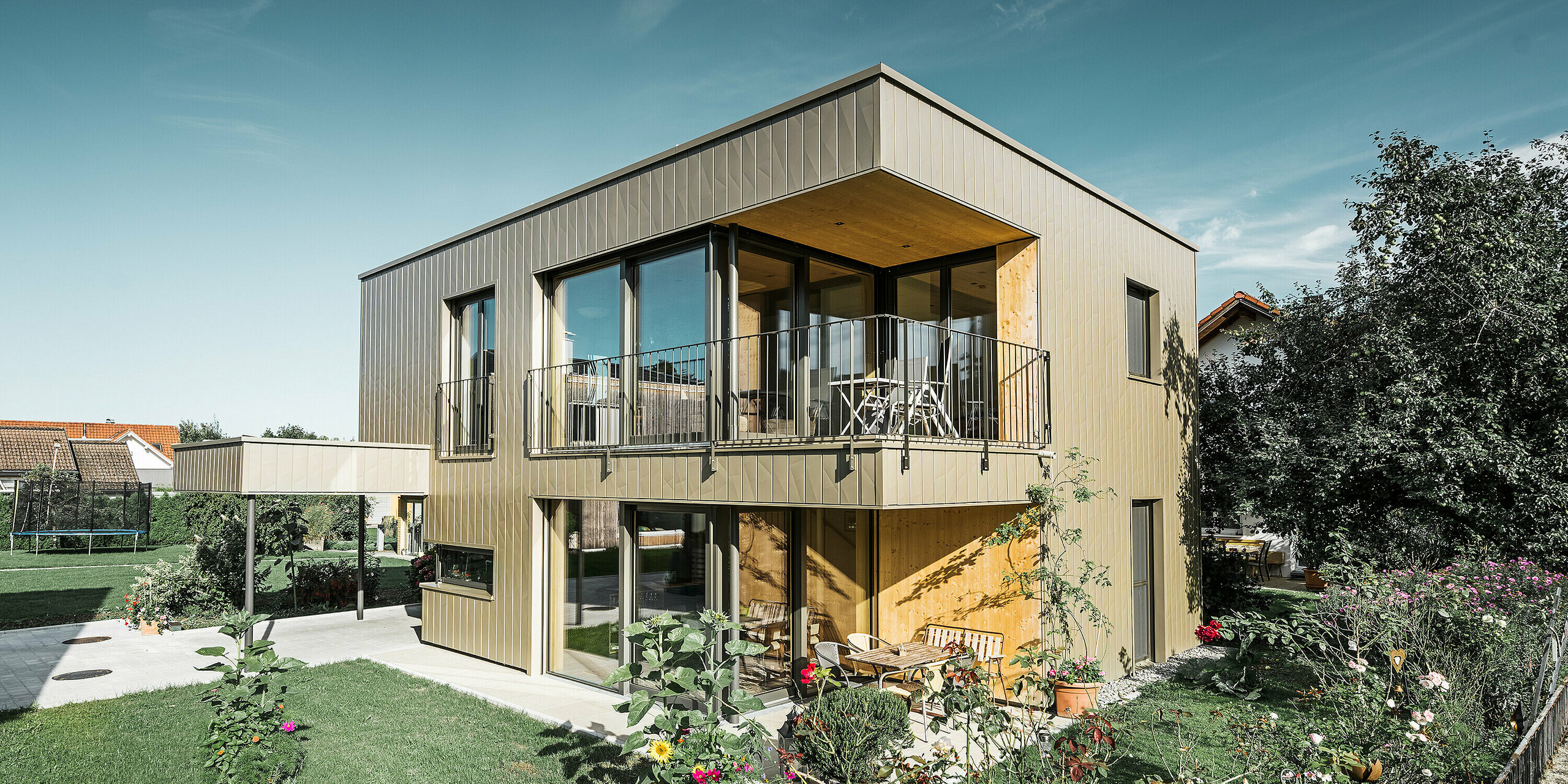 Moderner Neubau mit Flachdach und großen Fensterflächen; Der Großteil der Fassade ist mit vertikalen Alu-Fassaden-Paneelen von PREFA, Siding.X, in Bronze verkleidet. Man erhält auch einen Blick auf den rundum liegenden Garten.