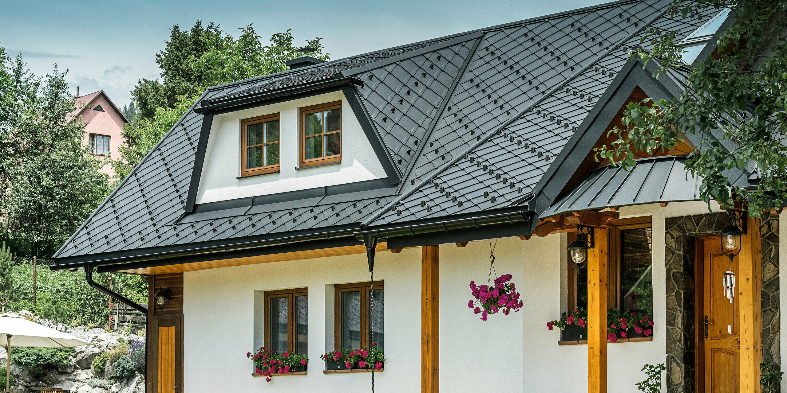 Vorderansicht eines traditionellen Einfamilienhauses in Stara Bystrica, Slowakei, mit einem Dach aus PREFA Dachrauten 29 × 29 in P.10 Schwarz. Das Dach mit dem einprägsamen Rautenlook umfasst Trapezgauben und Vordächer. Die klare Linienführung und die sorgfältig platzierten Fenstern verleihen dem Gebäude ein ansprechendes Erscheinungsbild. Blumenarrangements und eine gepflegte Gartengestaltung runden das Gesamtbild ab.