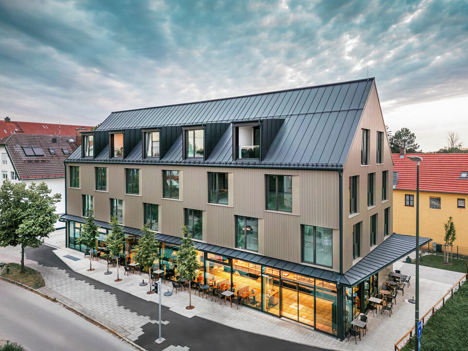 Das Bild zeigt ein modernes Gebäude mit einer Fassadenverkleidung aus Prefalz in der Farbe Bronze und einem Satteldach, das ebenfalls mit Prefalz in Schwarzgrau gedeckt ist. Die große Fensterfront im Erdgeschoss wird von Bäumen umrahmt und schafft eine einladende Atmosphäre für die darunter liegende Gastronomiefläche. Die Kombination aus den hochwertigen Prefalz-Materialien und der modernen Architektur verleiht dem Gebäude ein elegantes und zeitgemäßes Erscheinungsbild.