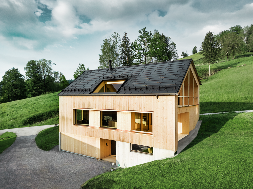 Ein zeitgenössisches Einfamilienhaus mit einer natürlichen Holzverkleidung fügt sich harmonisch in eine hügelige Landschaft ein. Das geneigte Dach ist mit schwarzen PREFA Solardachplatten und den Dachplatten R.16 ausgestattet, was einen modernen Kontrast zum warmen Holz bietet. Eine geschwungene Zufahrtsstraße und ein sattgrüner Rasen umgeben das Haus. 