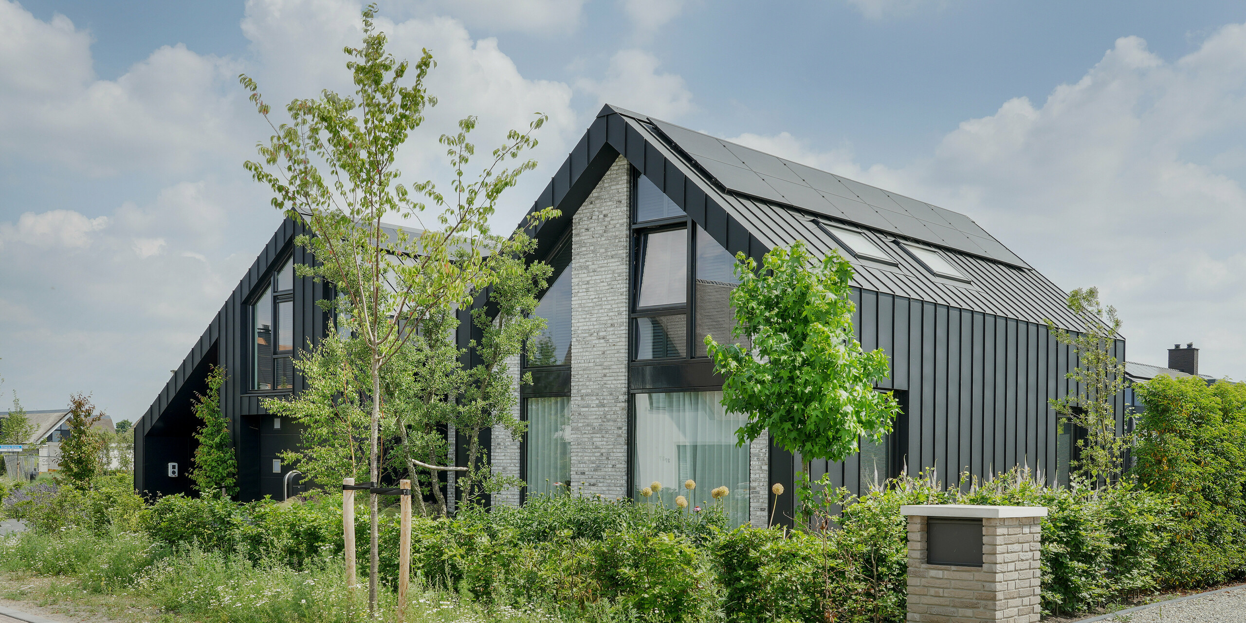 Moderne Wohnarchitektur in Veenendaal, Niederlande, geprägt durch schwarzes Aluminium von PREFA auf dem Dach und Teilen der Fassade. Das Haus mit seinen spitzen Giebeln und dem Stufendach hat eine starke geometrische Form. Die Verwendung von hellgrauem Naturstein als Teil der Fassade bietet einen subtilen Kontrast zu den dunklen Stehfalzziegeln. Das Haus ist von einer natürlichen, wilden Gartenlandschaft und gepflasterten Wegen umgeben, die eine Verbindung zwischen der modernen Architektur und der natürlichen Umgebung schaffen.