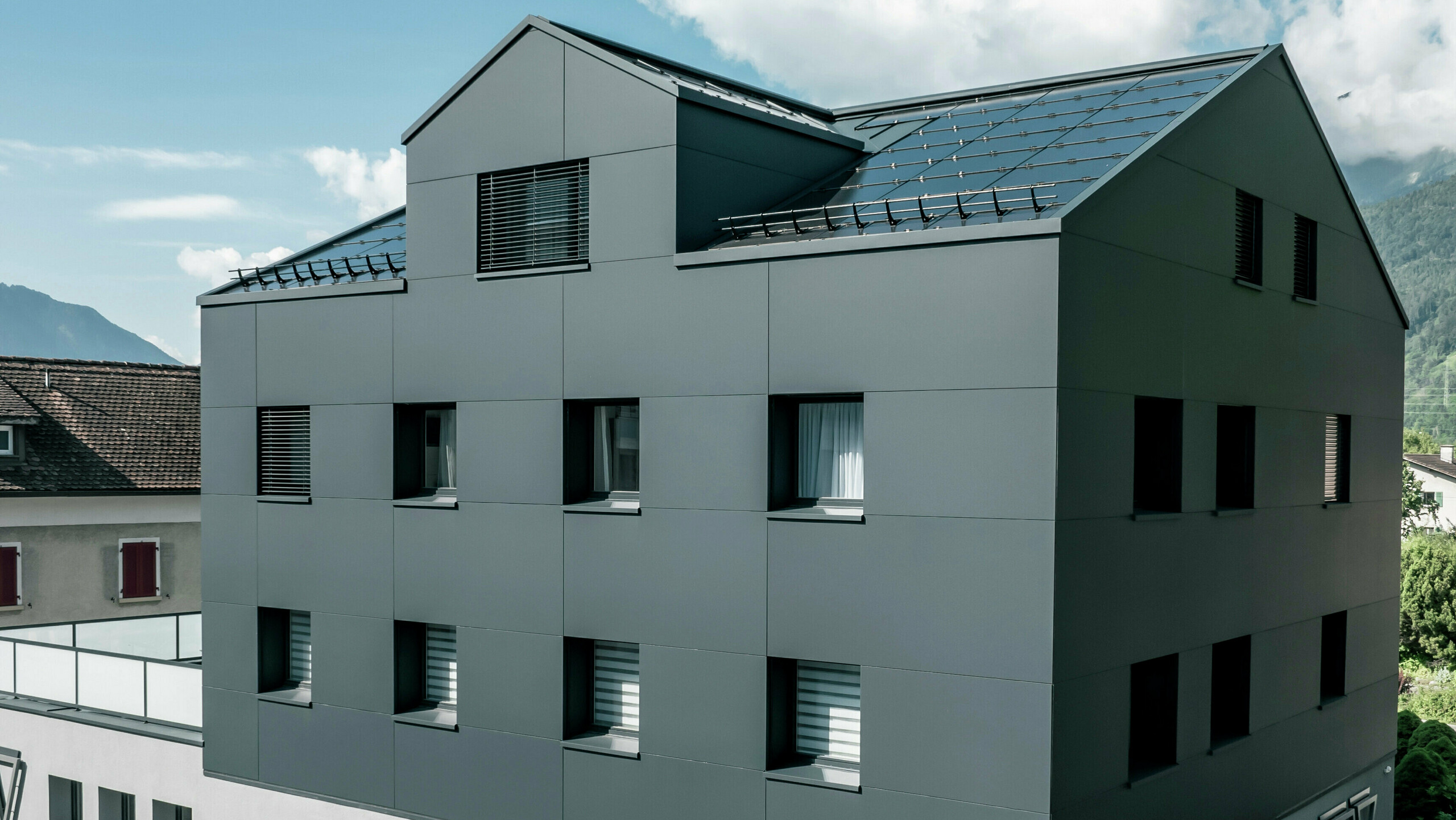 Modernisiertes Wohn- und Geschäftsgebäude nach der Fassadensanierung mit PREFABOND Aluminium Verbundplatten in Anthrazit. Die klare, elegante Linienführung der neuen Fassade verleiht dem Gebäude ein modernes Erscheinungsbild. Im Erdgeschoss befindet sich das Architekturbüro Voser. Die Umgebung mit Bäumen und Bergen rundet die harmonische Optik ab.