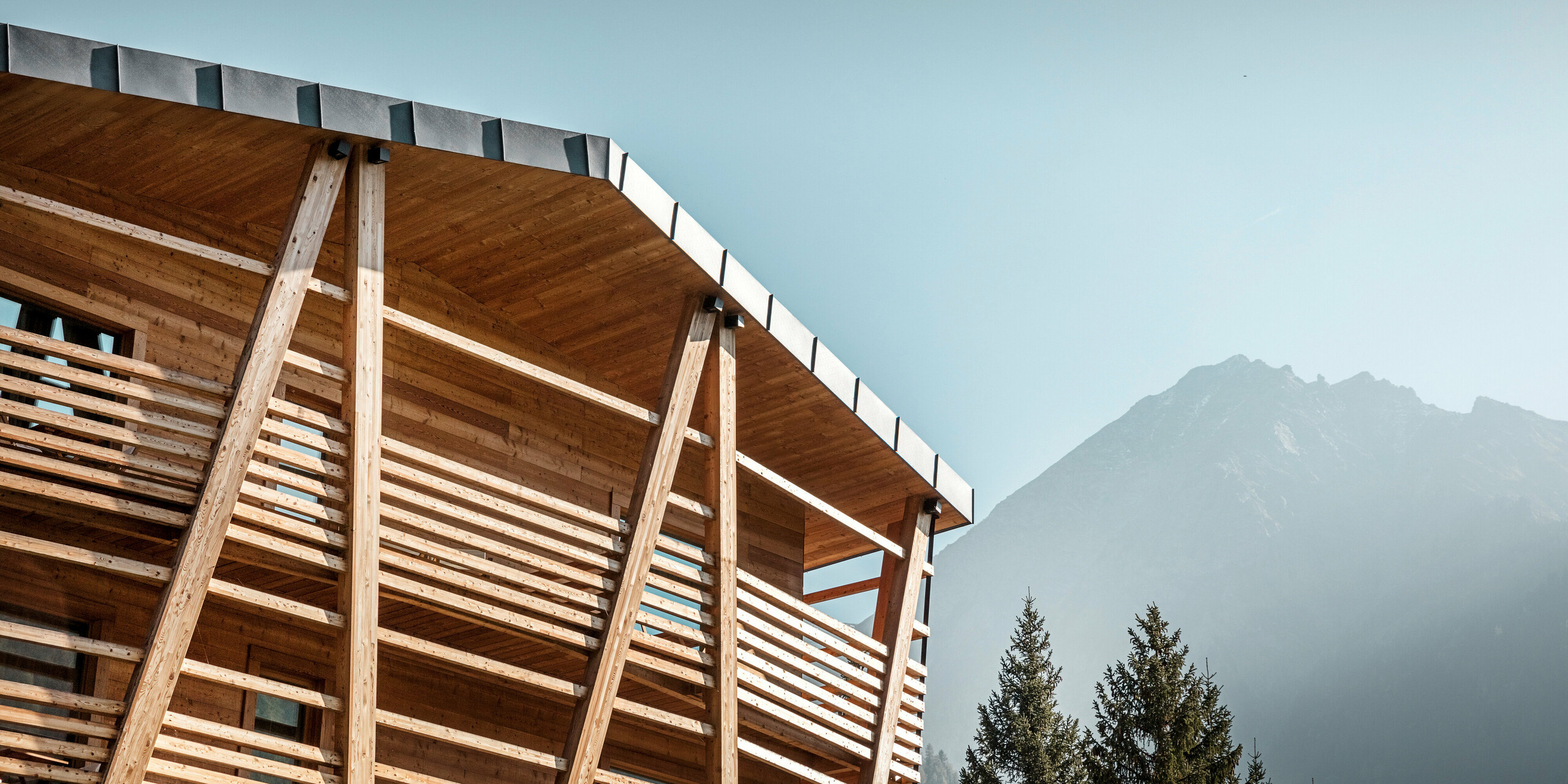 Nahaufnahme des Hotels 'Au Charmant Petit Lac', das mit seinen sichtbaren Holzstrukturen und dem darüber liegenden PREFALZ Aluminiumdach in P.10 Steingrau vor einer beeindruckenden Bergkulisse steht. Die Dachkante zeigt die präzise Verarbeitung der Stehfalzdeckung, die für ihre Langlebigkeit und Widerstandsfähigkeit gegen die alpinen Wetterbedingungen bekannt ist. Der Kontrast zwischen der natürlichen Holzfassade und dem metallischen Glanz des Daches betont das umweltfreundliche Design und die anspruchsvolle Architektur des Gebäudes.