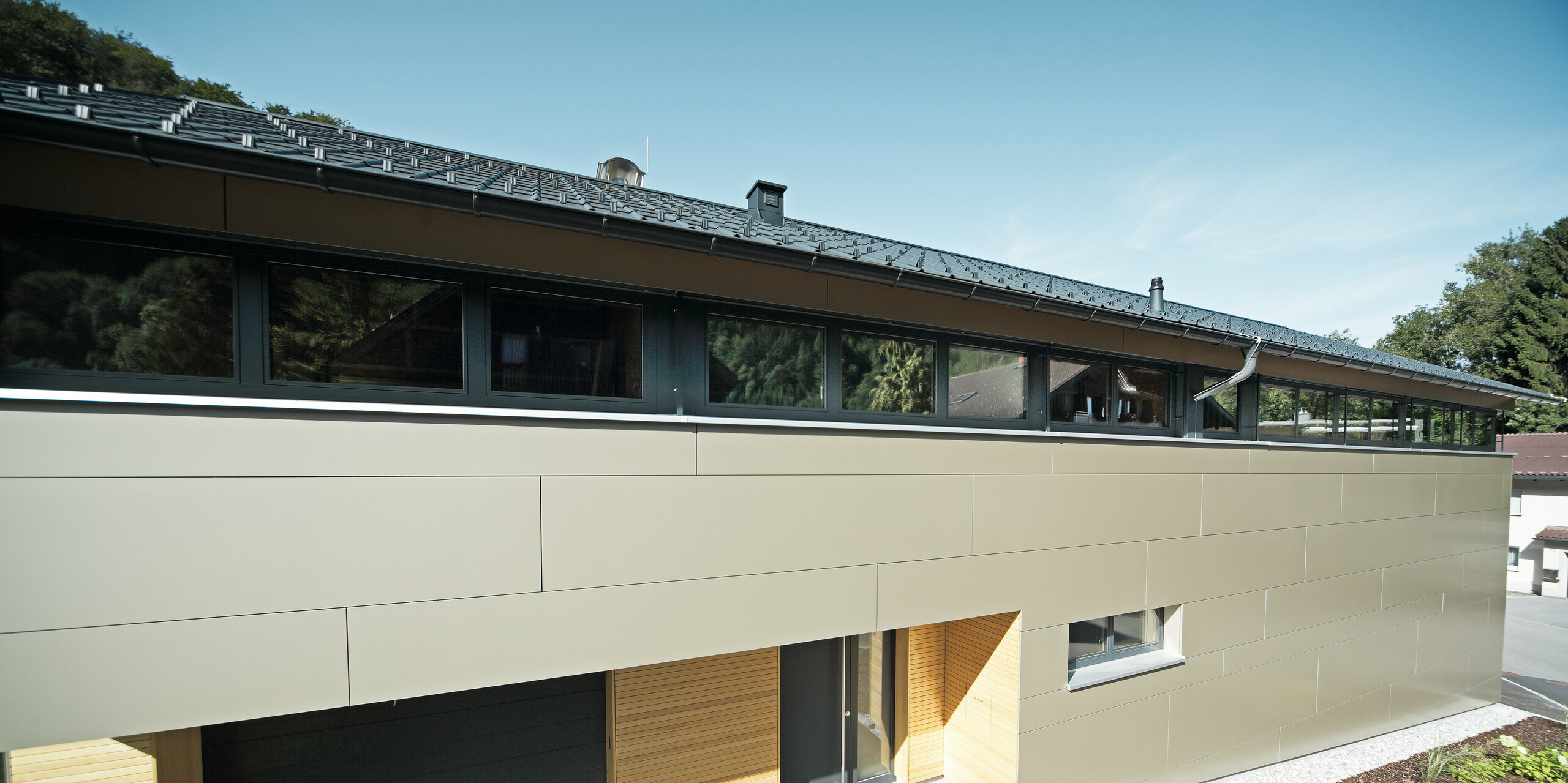 Frontansicht eines Einfamilienhauses in Hohenems, das mit PREFA Aluminium Verbundplatten in der Farbe Bronzemetallic verkleidet ist. Die horizontale Linienführung der Fassadenplatten schafft eine visuelle Breite und Struktur, während die Holzelemente im Eingangsbereich einen natürlichen Kontrast setzen. Das Dach ist mit PREFA Dachplatten in P.10 Anthrazit gedeckt, die perfekt mit der Fassade harmonieren und das moderne Design des Hauses unterstreichen.