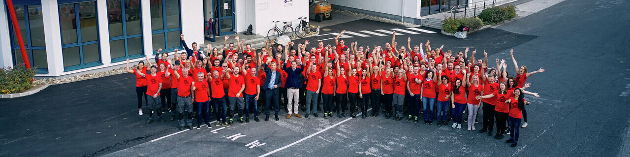 PREFA MitarbeiterInnen mit rotem PREFA T-Shirt stehen in einem Halbkreis vor dem Bürogebäude in Marktl.