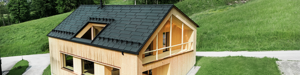 Ein modernes, zweistöckiges Haus mit einer Fassade aus hellem Holz steht in einer idyllischen, grünen Landschaft. Das schräge Dach des Hauses ist mit schwarzen PREFA Solardachplatten und R.16 Dachplatten versehen. Vor dem Haus befindet sich ein gepflegter Rasen, und eine geschwungene Schotterstraße führt zum Haus. Im Hintergrund erstrecken sich sanfte grüne Hügel mit Bäumen.